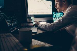 Male stock market trader using technologies while analyzing graphs and charts in the night office photo