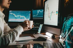 Male trader holding coffee cup while analyzing stock market data on multiple computers at night office photo