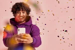 Joyful Indian man using smart phone while confetti flying around against pink background photo