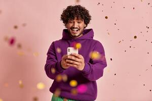 Excited Indian man using smart phone while confetti flying around against pink background photo