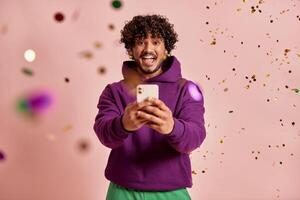 Happy Indian man using smart phone while confetti flying around against pink background photo