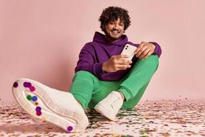 Handsome Indian man using smart phone while sitting on confetti against pink background photo