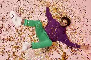 Top view of relaxed Indian man gesturing and smiling while lying on colorful confetti photo