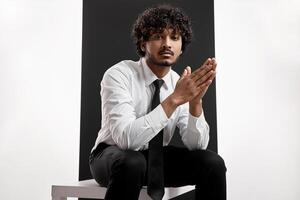 Handsome Indian man in shirt and tie looking at camera while sitting against black background photo