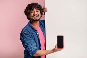 Happy Indian man showing smart phone while leaning on copy space against pink background photo
