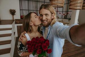 Handsome young man making selfie while his girlfriend kissing him and holding flower bouquet at home photo