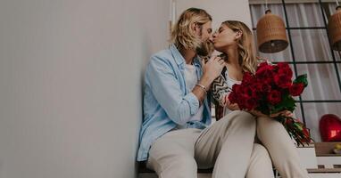 Young loving couple embracing and kissing at home while woman holding bouquet of red roses photo