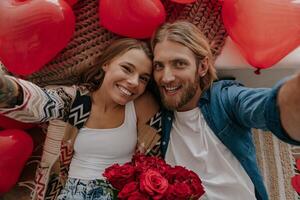 Top view of happy loving couple holding flowers and making selfie with red heart shape balloons around them photo