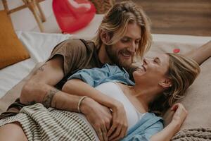 Relaxed loving couple embracing and smiling while lying in bed surrounded with red heart shape balloons photo