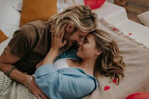 Young loving couple embracing and kissing while lying in bed surrounded with red heart shape balloons photo