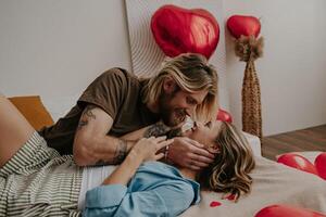 Relaxed loving couple embracing and smiling while lying in bed surrounded with red heart shape balloons photo