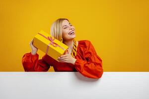 Curious young woman holding a gift box near face and smiling against yellow background photo