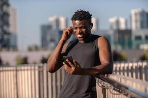 Cheerful African man in sportswear using smart phone and adjusting headphones while resting after training photo