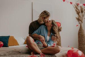 Romantic loving couple sitting on bed and kissing while red heart shape confetti flying around photo