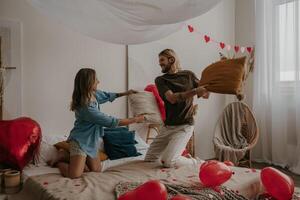 Smiling young loving couple having a pillow fight in bed surrounded with red heart shape balloons photo