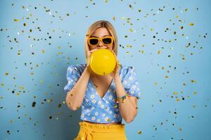 Happy young woman blowing up a balloon while confetti flying around against blue background photo