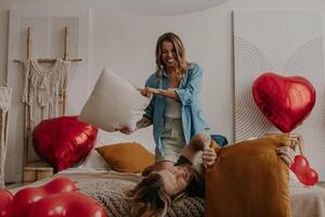 Joyful young loving couple having a pillow fight in bed surrounded with red heart shape balloons photo