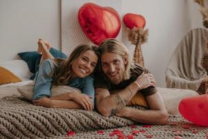 Smiling young loving couple lying in bed surrounded with red heart shape balloons and confetti at home photo
