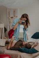 Beautiful young woman photographing her boyfriend with retro camera while both relaxing in bed at home photo
