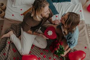 Top view of romantic young man giving a red heart shape gift box to his girlfriend while sitting on bed together photo