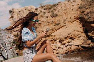 Attractive young woman using smart phone and smiling while sitting seaside photo