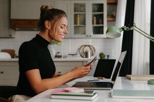 Smiling young woman sitting at the desk and using smart phone while working at home photo