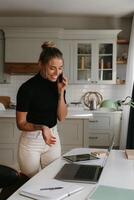 Confident young woman talking on mobile phone and looking at laptop while working at home photo