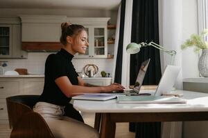 Side view of attractive young woman sitting at the desk and using laptop while working at home photo