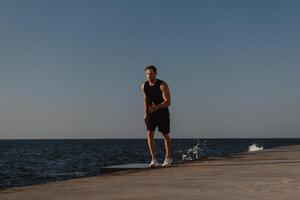 Confident athletic man in sports clothing running by the quay with the sea on background photo
