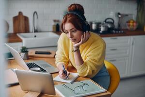 Beautiful woman in headphones making notes and looking at laptop while sitting at the kitchen counter photo