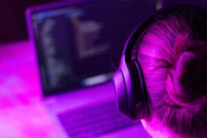 woman software developer in modern wireless headphones against laptop screen with software code, neon light, suitable for software development , IT education topics photo