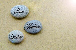 Top view of Love, Kindness and Devotion Joy text engraved on stones with beach sand background. photo