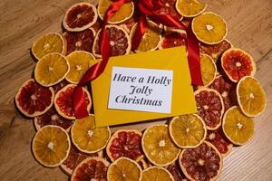 Christmas handmade wreath made of dried slices of orange and grapefruit with yellow envelope and paper note with HAVE A HOLLY JOLLY CHRISTMAS text. Winter holiday celebration greeting card. top view photo