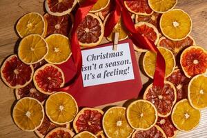 Christmas handmade wreath made of dried slices of orange and grapefruit with red envelope and paper note with CHRISTMAS ISN'T A SEASON IT'S A FEELING text. Winter holiday celebration greeting card. top view photo