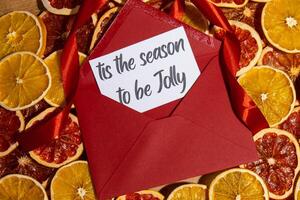 IT'S THE SEASON TO BE JOLLY text on postcard with red envelope on dried slices of grapefruit and oranges in Christmas wreath. Zero waste eco-friendly Christmas. Sustainable greeting card decoration photo