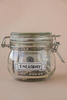 Saving Money In Glass Jar filled with Dollars banknotes. EMERGENCY transcription in front of jar. Managing personal finances extra income for future insecurity. Beige background photo