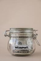 Saving Money In Glass Jar filled with Dollars banknotes. RETIREMENT transcription in front of jar. Managing personal finances extra income for future insecurity. Beige background photo