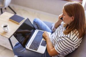 Happy young woman using mobile phone while sitting on sofa at home with laptop photo