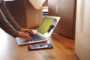 Female hands are typing on a laptop on the parquet floor. There are a telephone nearby and a lot of cardboard boxes with things. The concept of moving from an apartment or house. photo