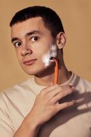 A close-up portrait of a teenager with foam on his cheek and holding a razor. A handsome guy in a T-shirt is shaving. Hygiene and skin care in adolescence. photo