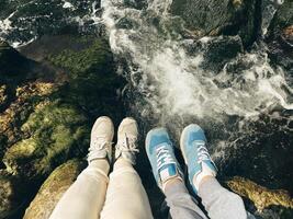 Feet in sneakers on the rocks, selfie over a fast river in the background. Top view of female and male legs over bubbling water. Tourists on a hike. photo