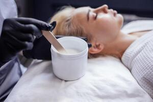 Close up of jar with scrub to cleanse the skin. cosmetologist hands in black gloves hold wooden spatula for applying cream. photo