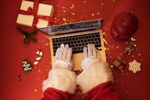 Papa Noel claus con ordenador portátil. manos en blanco guantes mecanografía en un cuaderno teclado. regalo cajas, Navidad galletas, linterna y abeto rama en rojo antecedentes. foto