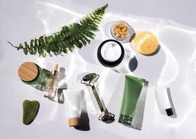 A set of mockups of ecological cosmetics with a fern leaf. Various cream tubes, a bottle of serum, capsules, a sponge, a jade facial massager and gouache scraper. photo