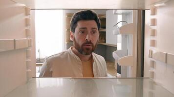 Hungry Caucasian bachelor man looking for food in empty fridge finding no meal products feeling sad disappointed poor unemployment need delivery service POV point of view from inside refrigerator photo