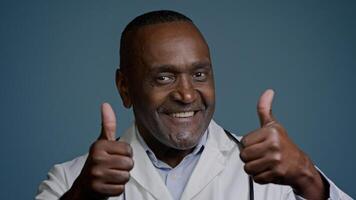 Portrait smiling African American man doctor in white coat showing thumbs up in studio blue background looking at camera gesture approval recommendation medical services health worker show like sign photo