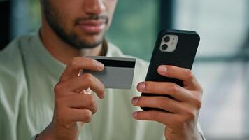 African American bearded man indoors unrecognizable male customer holding credit card and smartphone sitting at home office using easy mobile payment app service making purchase buying in online store photo