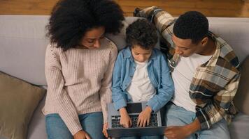 Top view African American family married couple teaching little boy child son kid offspring typing using tech laptop notebook pc device discuss speaking tell online education indoors living room couch photo