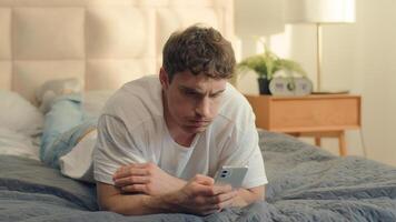 Caucasian unhappy man lying on bed user smartphone mobile phone cellphone frowning breakdown spam male guy crash app device worry banking confused displeased social media lost connection network fraud photo