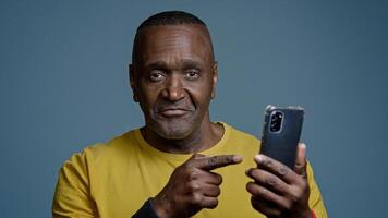 Mature man standing in studio on gray background holding mobile phone browsing social network sports athlete watching on internet physical exercise pointing to smartphone screen nods head approves photo